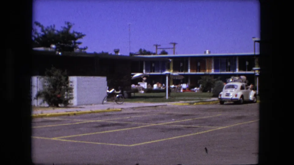 1976 Fort Wayne motel or roadside lodging view behind a parking lot, with a white Volkswagen Beetle carrying roof luggage and people gathered on the lawn.