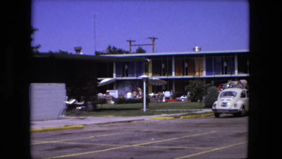 Fort Wayne 1976 parking lot looking toward a low motel-like building, lawn chairs or seating, a bicycle, distant people, and a white Volkswagen Beetle with luggage on top.