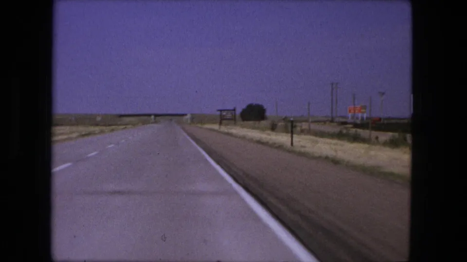 A straight road is filmed through a moving vehicle windshield, with flat roadside land and distant signs under a purple-tinged sky.