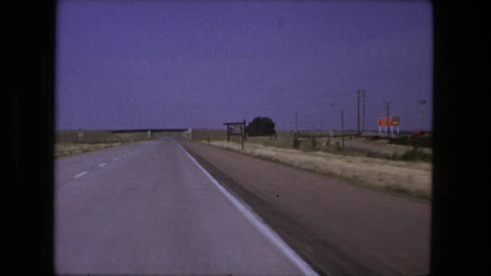 The windshield view continues along a straight highway with open land, roadside posts, and small distant signs in 1970s home-movie footage.