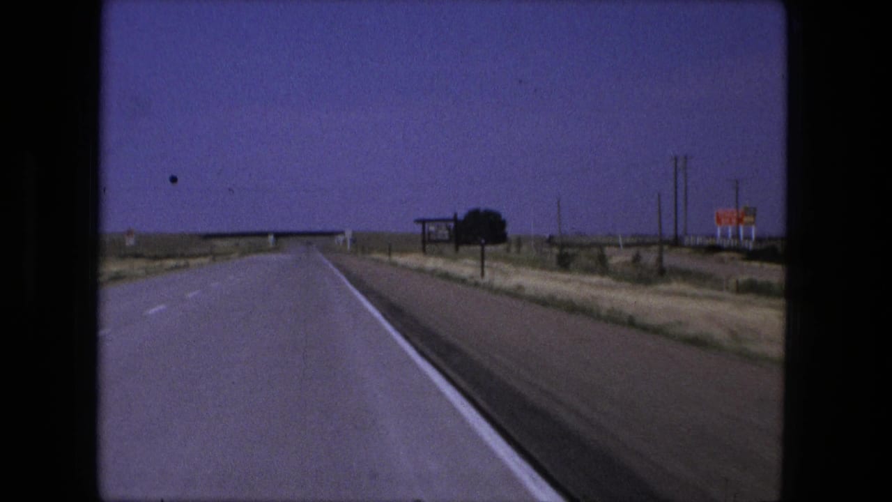 1976 8mm road-trip view near Fort Wayne, Indiana, looking down a rural highway toward signs and a distant bridge.