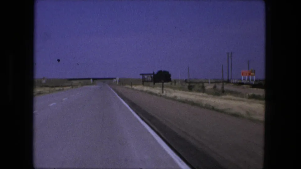 1976 8mm road-trip view near Fort Wayne, Indiana, looking down a rural highway toward signs and a distant bridge.