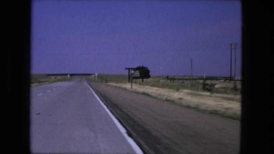 1976 home-movie frame from a vehicle on a rural Fort Wayne-area highway, showing the shoulder, sign, utility poles, and bridge approach.