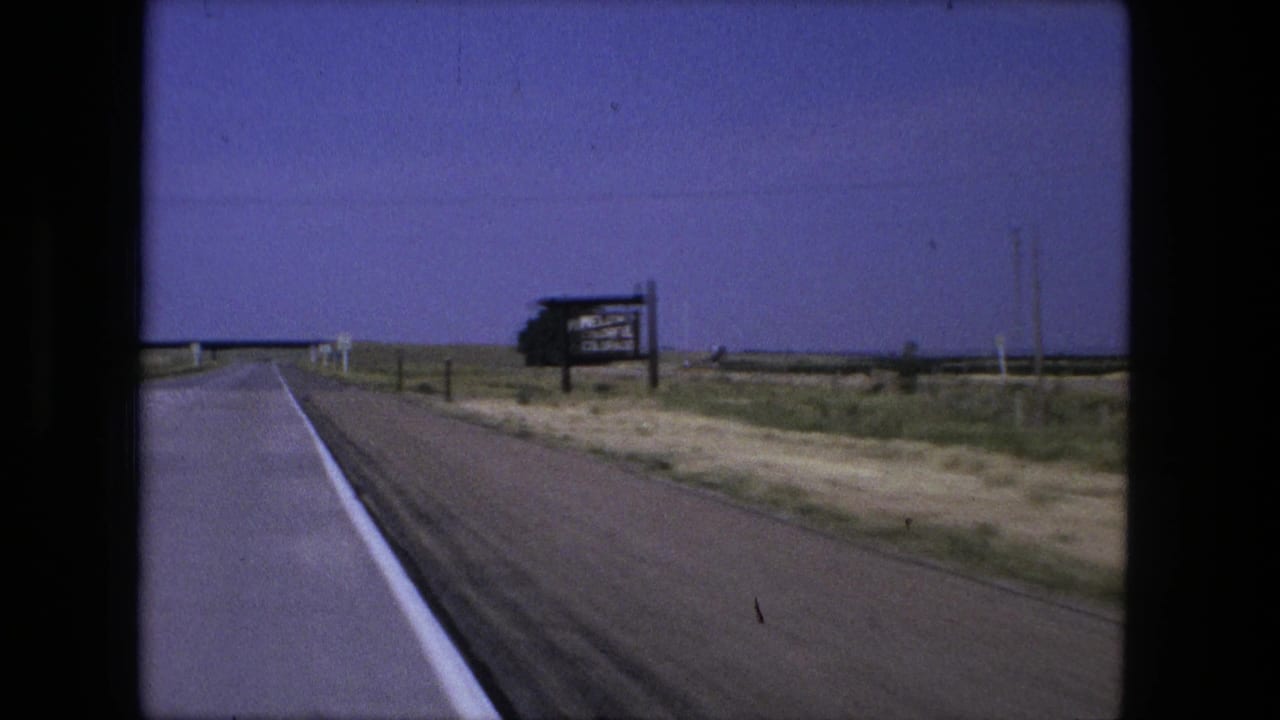 Rural road seen from a moving car with a distant roadside welcome sign and flat fields.