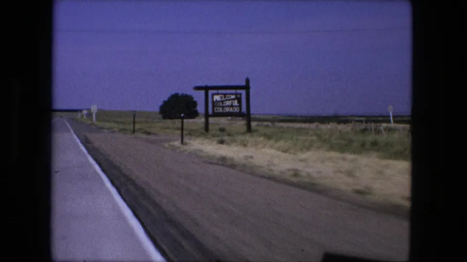 1976 8mm frame from a moving vehicle passing a Welcome to Colorful Colorado sign beside open rural land.