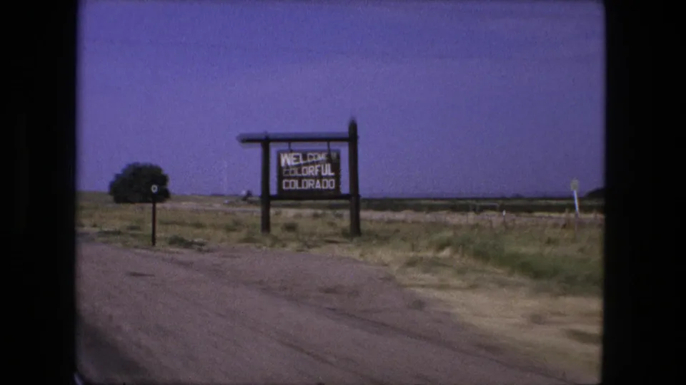 1976 8mm roadside view showing the Welcome to Colorful Colorado sign close to the car window, with dry fields behind it.