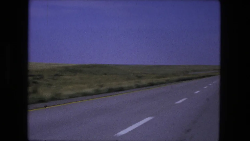 1976 8mm driving frame showing an open highway stretching through flat grassy fields after the roadside sign has passed.