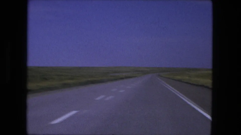 1976 8mm view of a straight rural highway near Fort Wayne, Indiana, with lane markings, grassy shoulders, and a distant horizon.