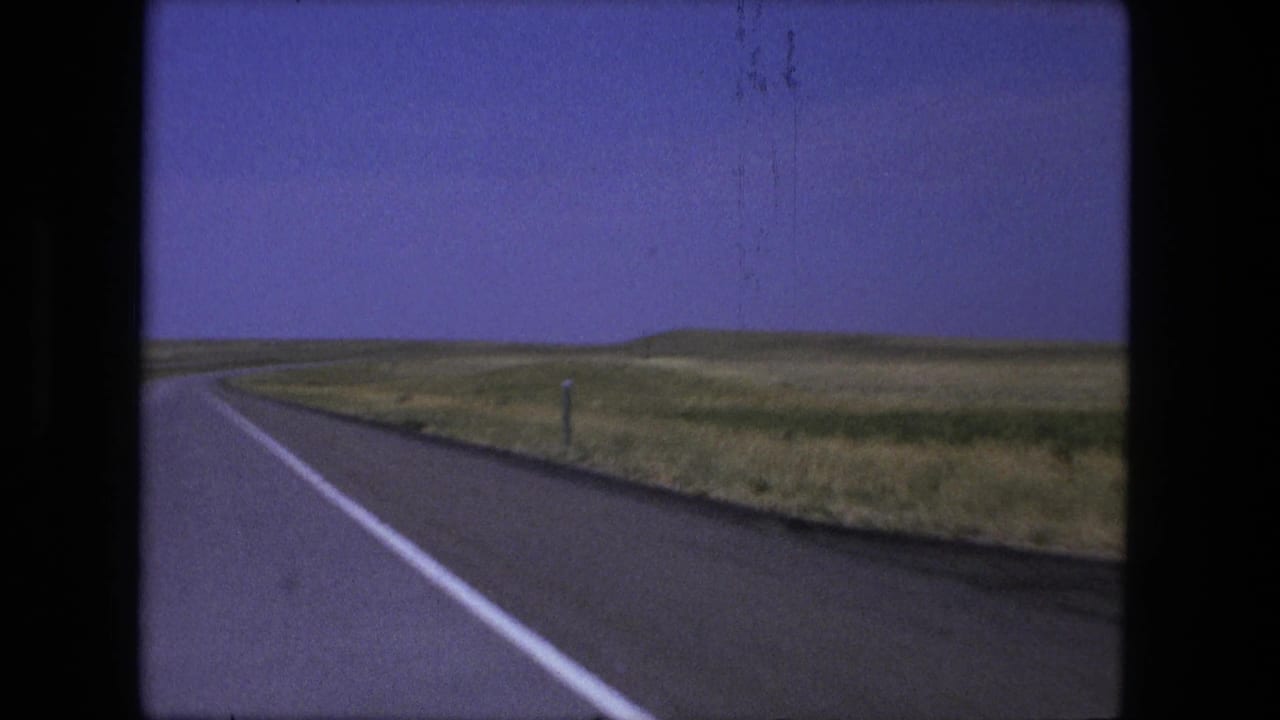 1976 8mm frame from a moving vehicle showing the highway shoulder, roadside post, open grassland, and low hills near Fort Wayne, Indiana.
