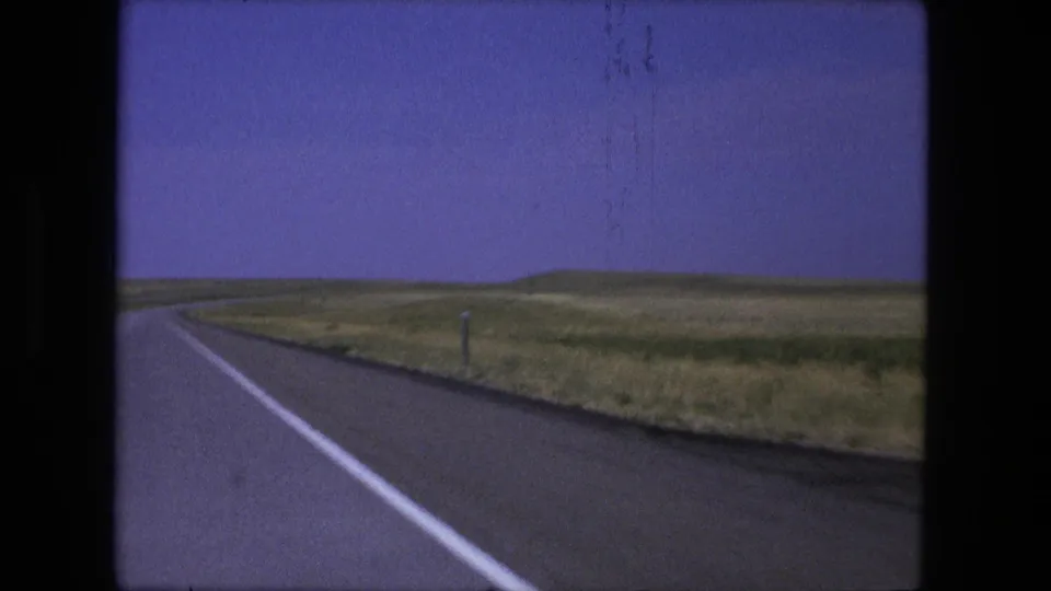 1976 8mm frame from a moving vehicle showing the highway shoulder, roadside post, open grassland, and low hills near Fort Wayne, Indiana.