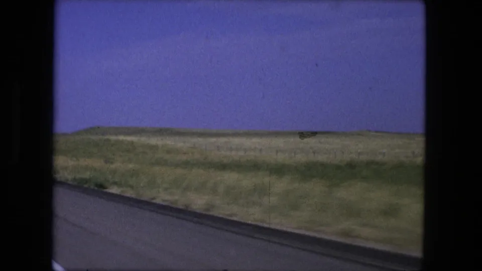 1976 8mm moving-vehicle view of pavement edge, roadside grass, low hills, and blue sky in rural Indiana.
