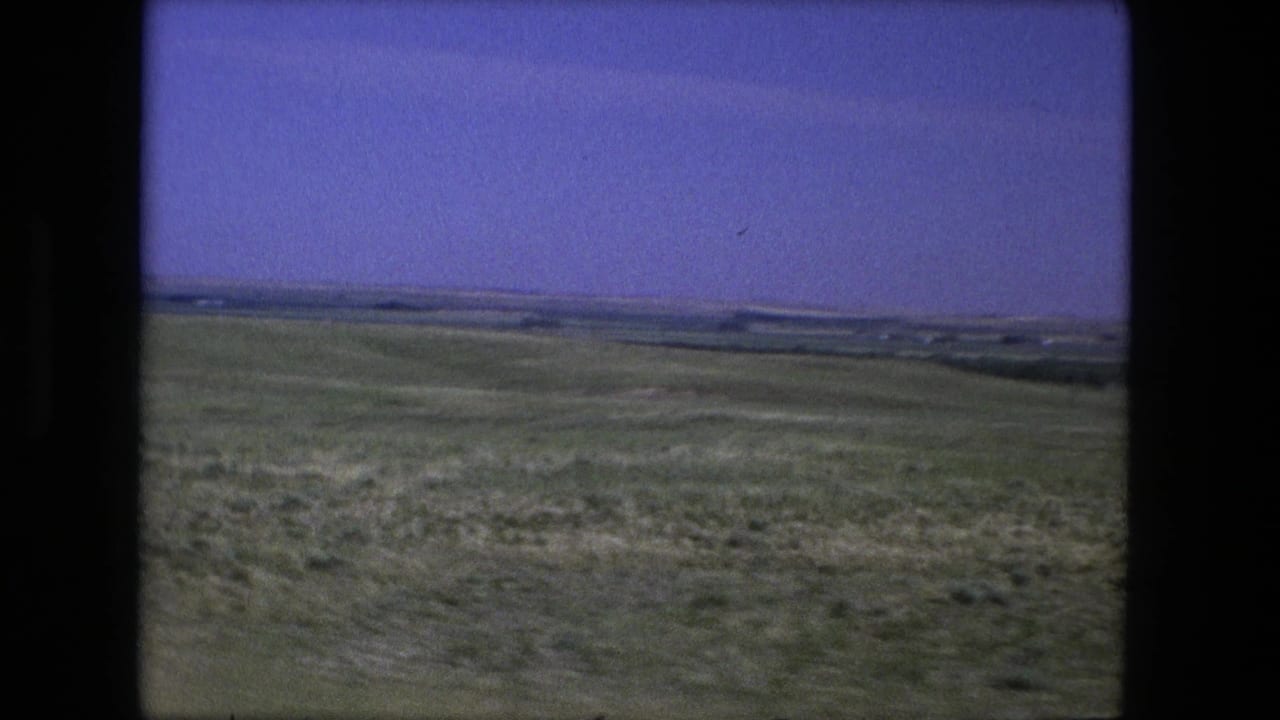 Grassy open plains and low hills beneath a blue sky, framed by dark film borders.
