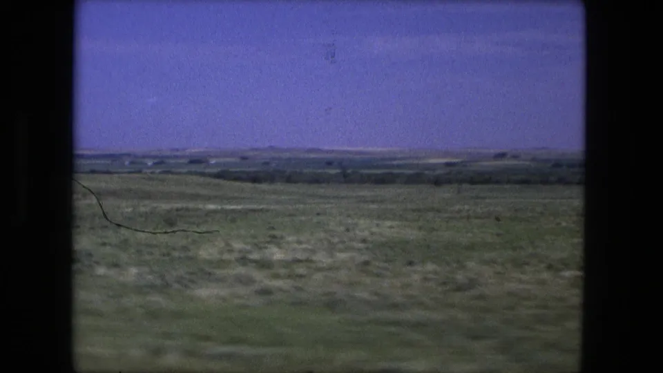 Rural Indiana landscape from 1976 8mm footage, showing rolling grassland, distant trees, and a hazy horizon.