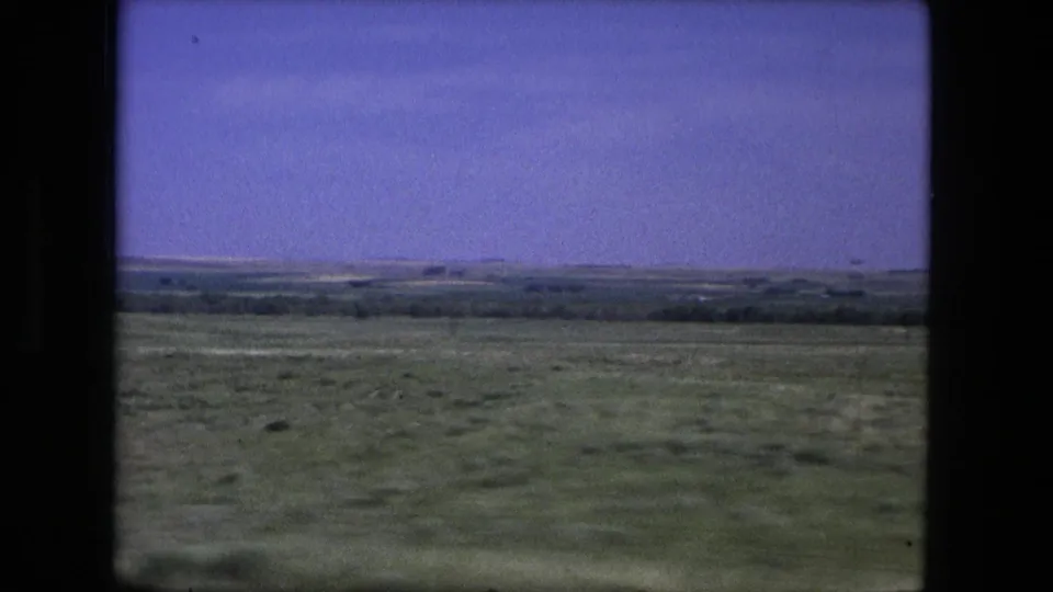 1976 home-movie frame of open countryside near Fort Wayne, with grassy foreground and distant low hills.