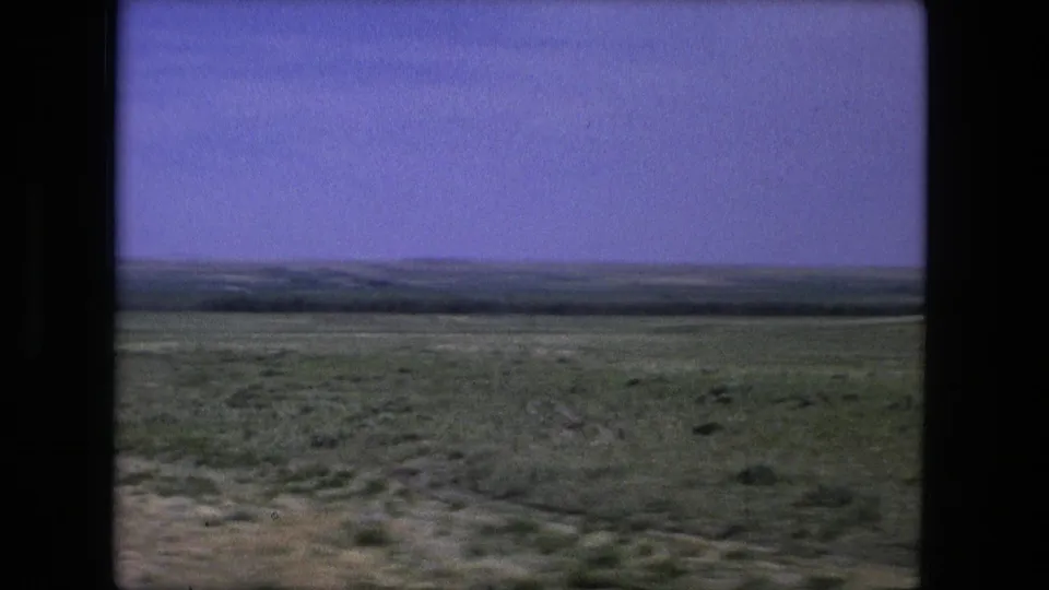 1976 8mm travel view of open grassland near Fort Wayne, with a faint ground track and distant horizon.