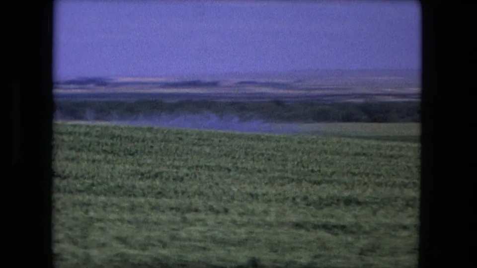 1976 8mm home-movie frame from the Fort Wayne, Indiana area showing green farmland, low horizon, and distant water or haze under a purple-blue sky.