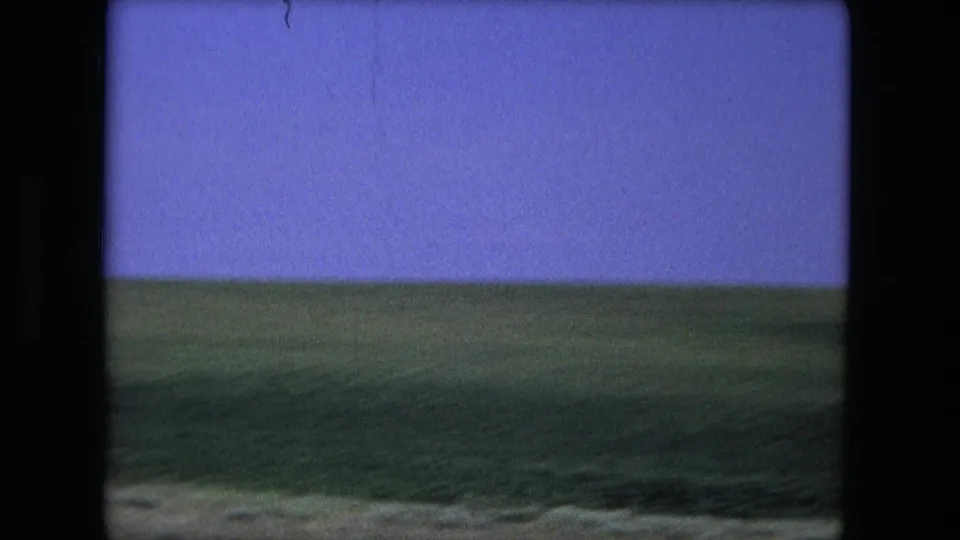 Motion-blurred 1976 8mm roadside view near Fort Wayne, Indiana, with green fields, low horizon, purple-blue sky, and film gate edges.