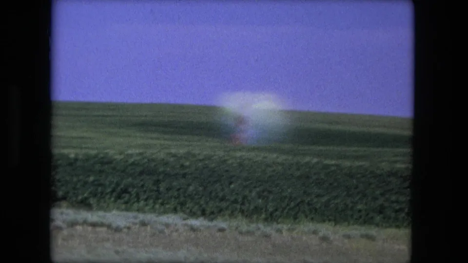 1976 8mm frame from the Fort Wayne, Indiana area showing green farmland, a purple-blue sky, and a bright indistinct form near the horizon.