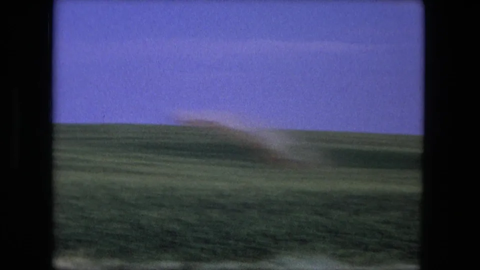 1976 8mm frame associated with Fort Wayne, Indiana, showing open grassland, a low horizon, and a blurred dust plume.