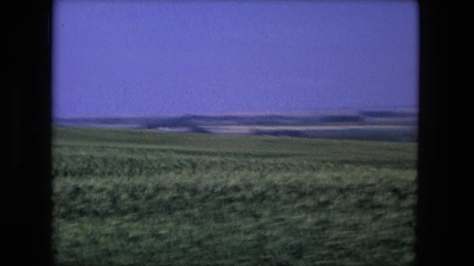 1976 8mm frame linked to Fort Wayne, Indiana, showing a broad green field and distant low landforms under a color-shifted sky.