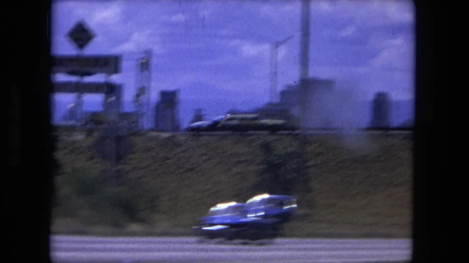 Roadside traffic passes an embankment and utility structures in 1976 Fort Wayne-area home-movie footage.