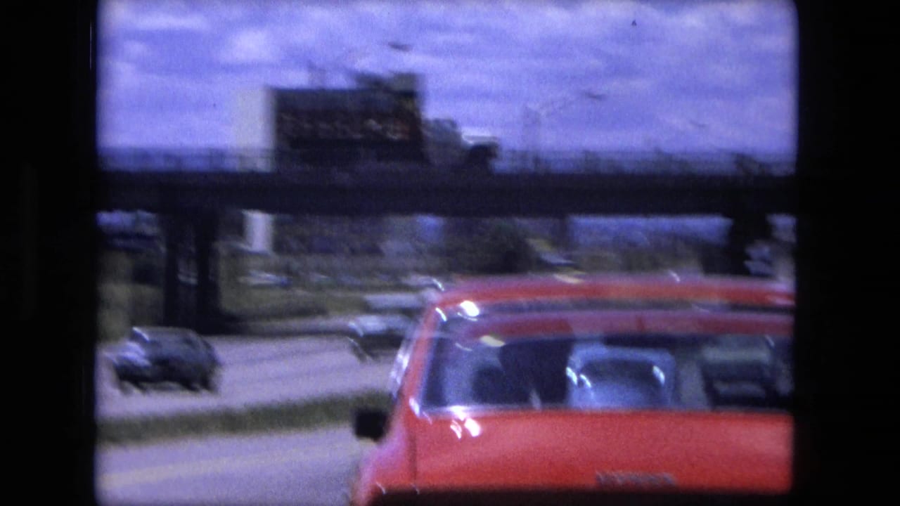 Red car on a highway seen through dark vehicle framing beneath an overpass.