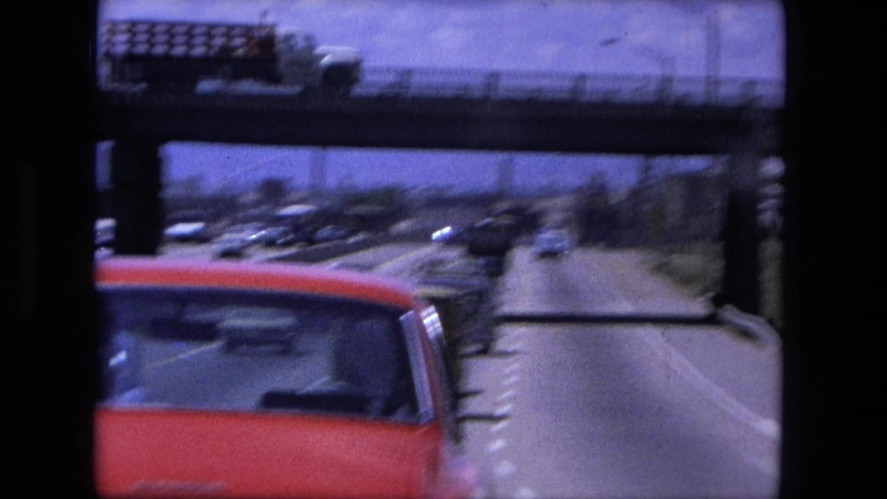 Red car close to the camera with traffic and an overpass ahead.