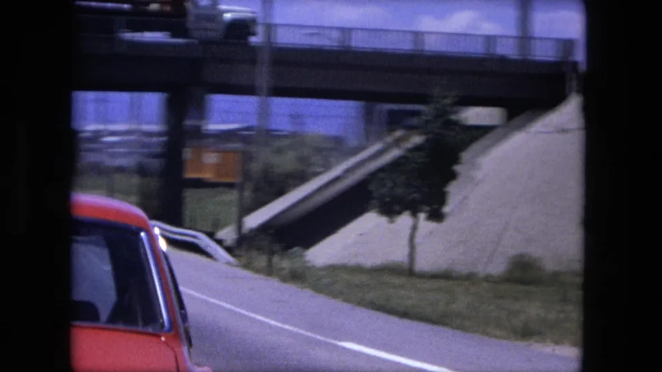 The red car slips partly out of frame as the camera passes a Fort Wayne overpass ramp and roadside embankment.