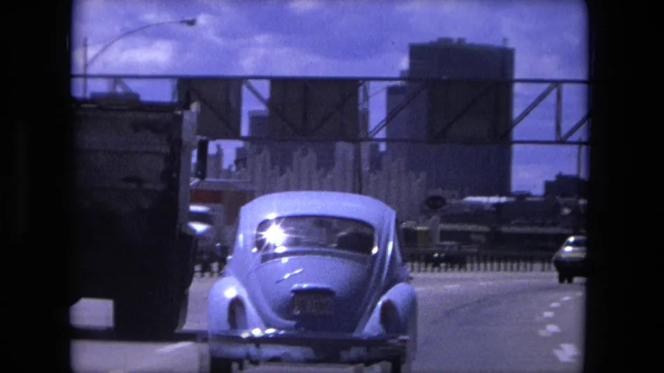 A white Volkswagen Beetle drives through Fort Wayne traffic in 1976, with bridge structures and other vehicles around it.