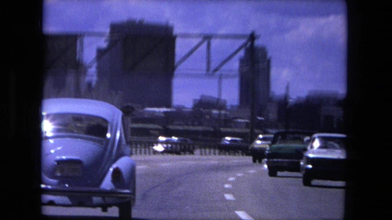 Cars travel along a highway toward distant city buildings under a bright sky.