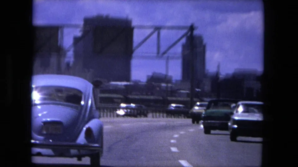 1976 Fort Wayne highway view with a light Volkswagen Beetle and other cars moving toward distant urban buildings.