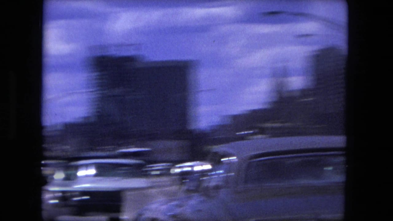 Blurred traffic passes on a roadway with blocky buildings in the background.