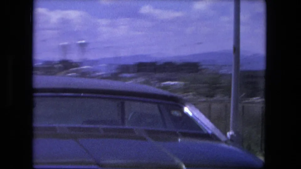 Passenger-side moving view in Fort Wayne, with a nearby car roof, roadside barrier, and distant low skyline visible through blue-tinted film.
