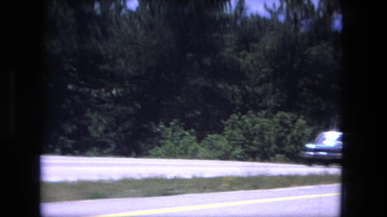1976 8mm road-view frame showing a paved highway edge, dense evergreens, blue sky, and dark window framing from a moving vehicle.