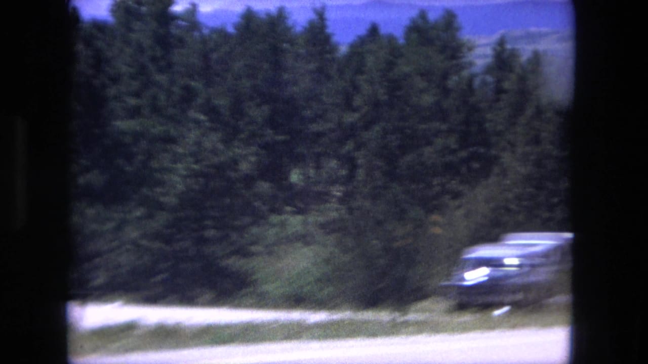 1976 8mm frame with a dark sedan blurred by motion beside a forested highway and heavy window-frame shadows.