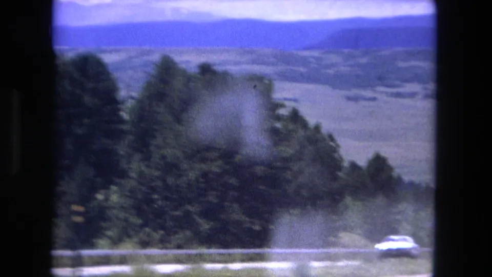 1976 8mm landscape frame showing distant blue mountains, open valley terrain, roadside trees, and a small car near the bottom edge.