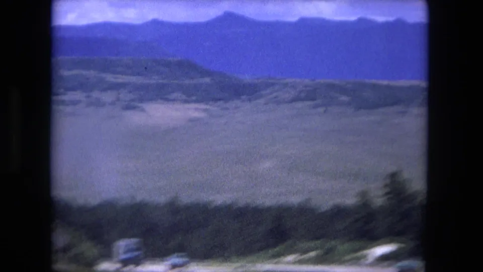 1976 8mm frame with layered mountain silhouettes, valley landforms, roadside blur, and dark vertical window framing.