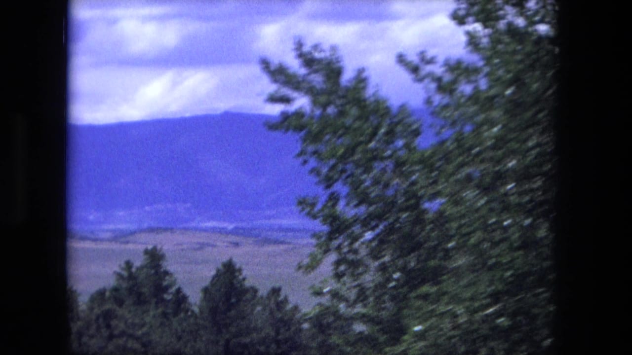 Cataloged 1976 home-movie frame showing roadside trees, distant hills, mountains, and cloud cover through a vehicle window.