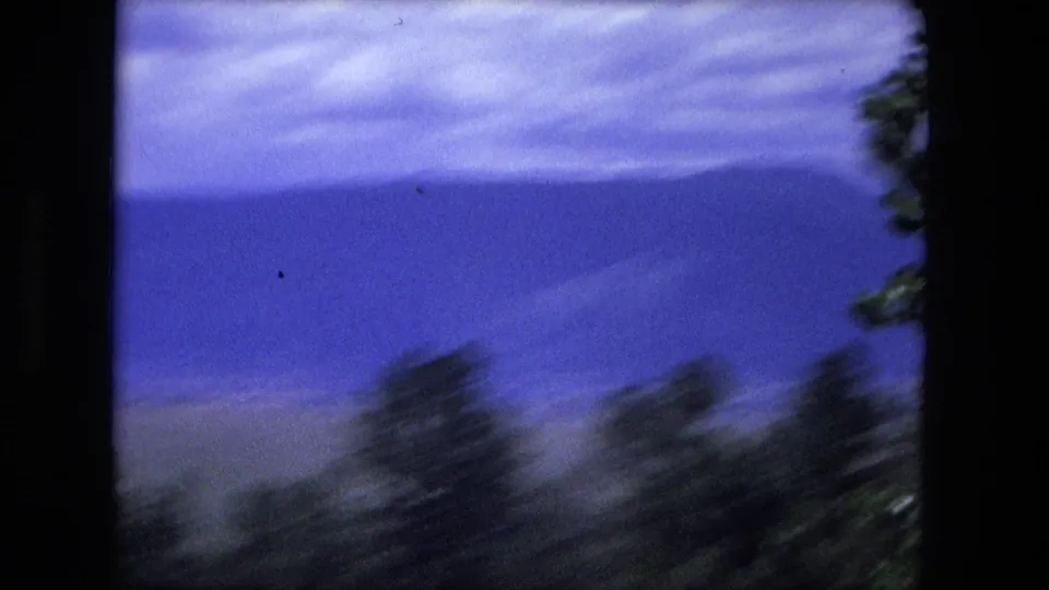 Moving-vehicle 8mm frame with motion-blurred trees, open hillside, a blue mountain ridge, and cloudy sky.