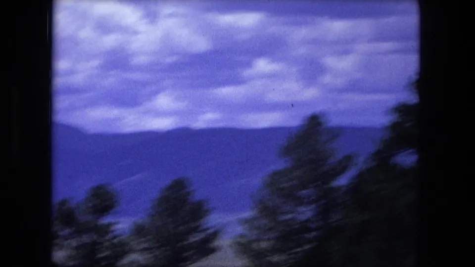 1976 cataloged 8mm frame with a dark window edge, soft trees, distant mountains, and textured cloud cover during travel.