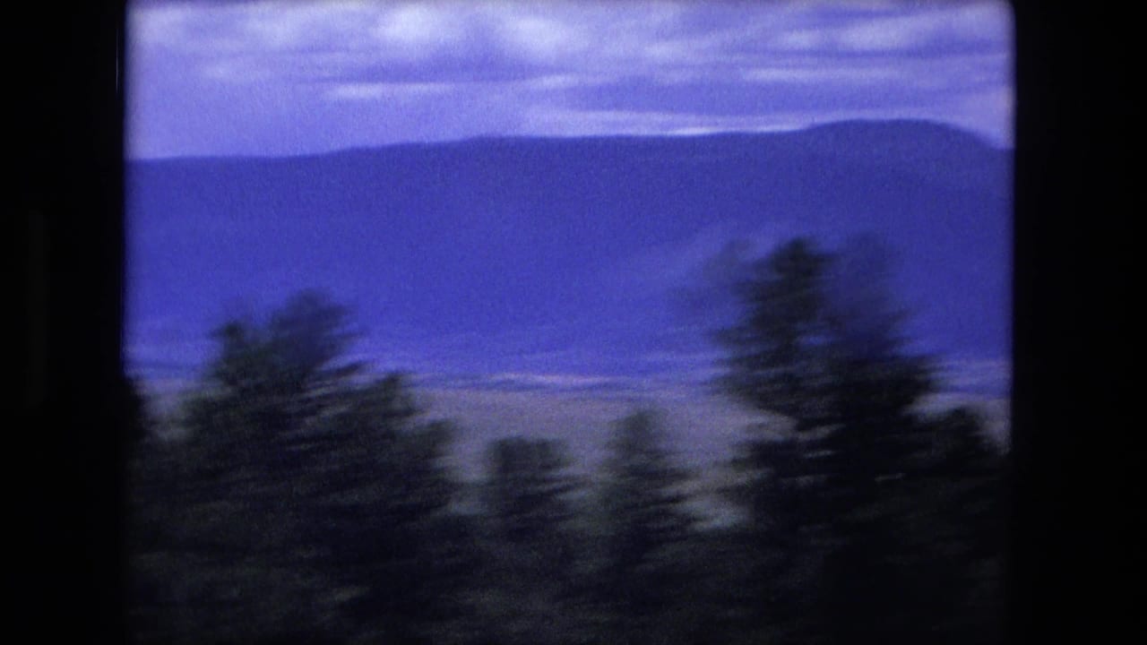 1976 8mm travel footage shows dark roadside trees blurred against distant mountain ridges under a blue-purple sky.