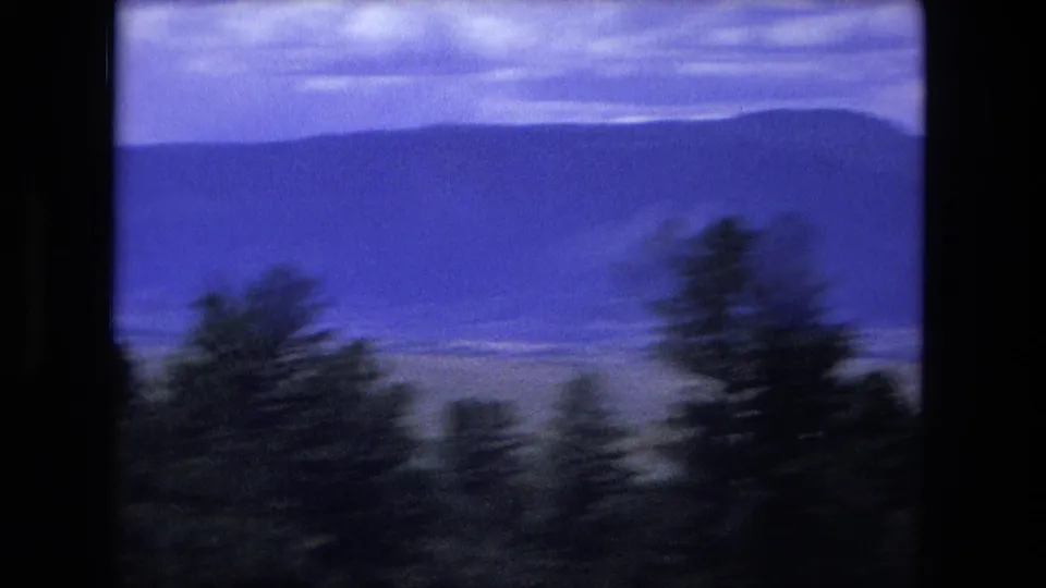 1976 8mm travel footage shows dark roadside trees blurred against distant mountain ridges under a blue-purple sky.