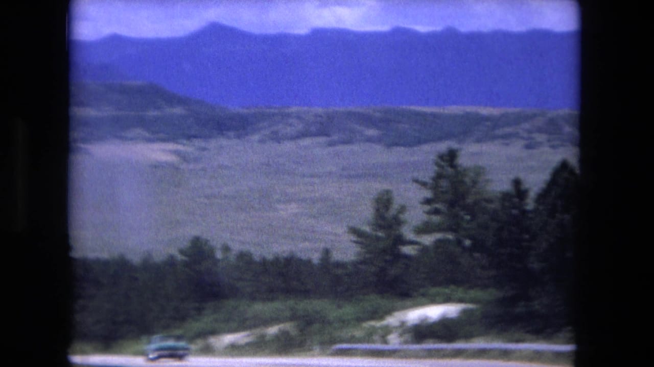 A small car travels along a road below open hills, dark trees, and distant mountains in 1976 8mm home-movie footage.