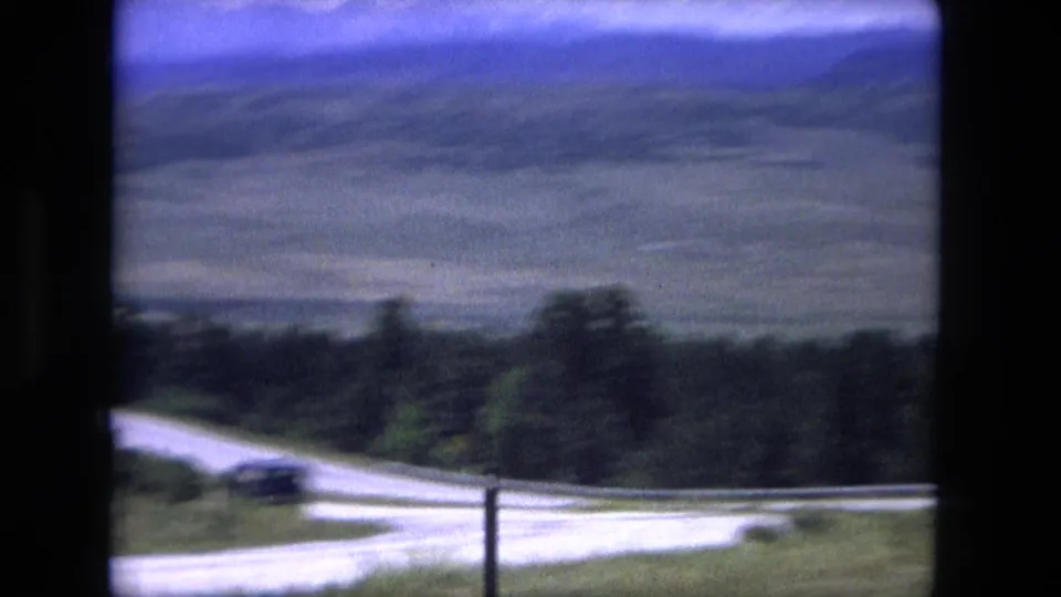 The moving camera looks over a curved pale road, roadside trees, rolling fields, and hazy distant mountains.