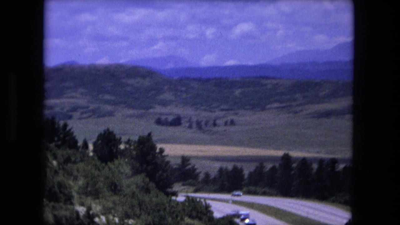 1976 8mm frame of a winding rural highway with small cars, trees, open hills, and distant mountains under a cloudy sky.