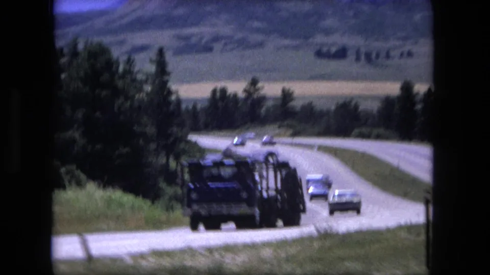 1976 8mm frame showing a large truck and several cars on a curving highway bordered by grass, trees, and distant hills.
