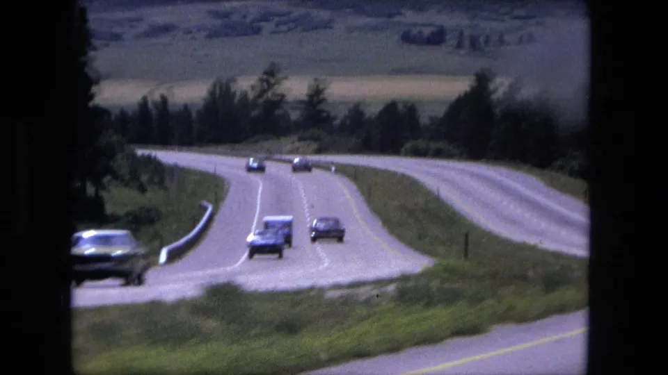 1976 8mm frame of cars on a divided highway winding through trees, open slopes, and distant hills.