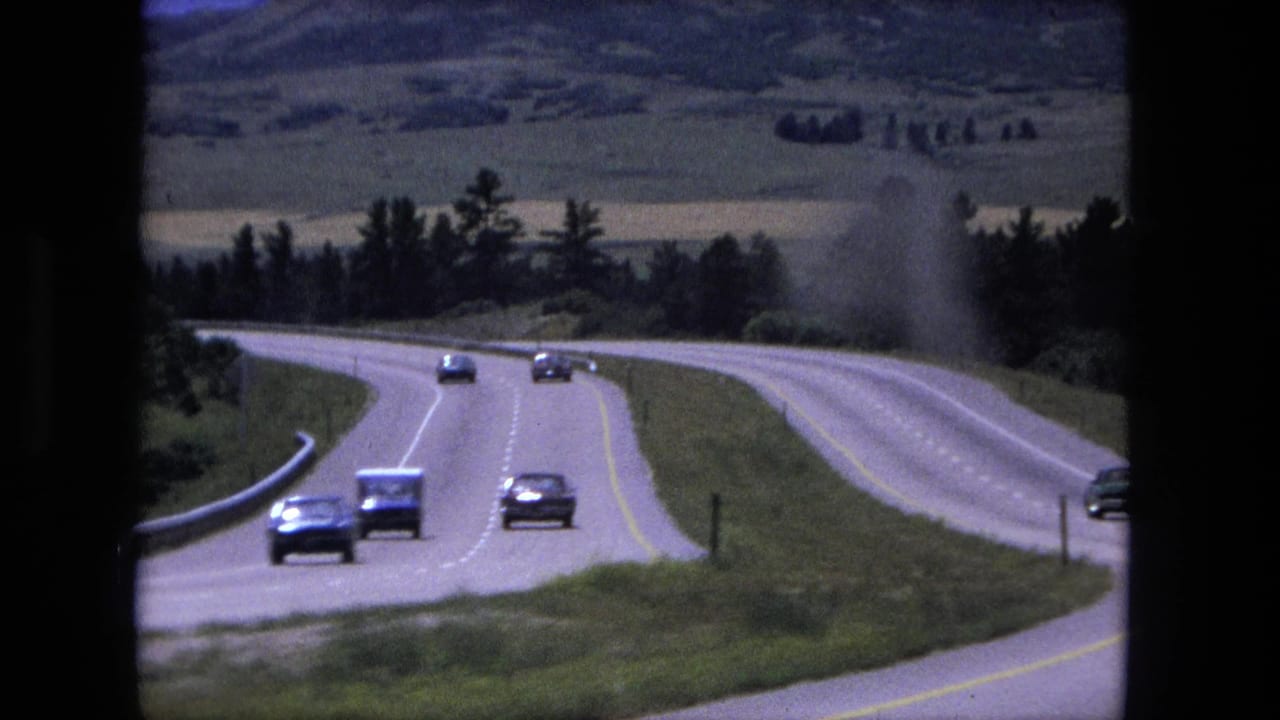Cars travel along a curving rural road bordered by grass, fields, and trees.
