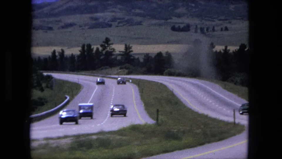 1976 8mm home-movie frame of cars on a winding rural road near Fort Wayne, Indiana, with fields and trees in the background.