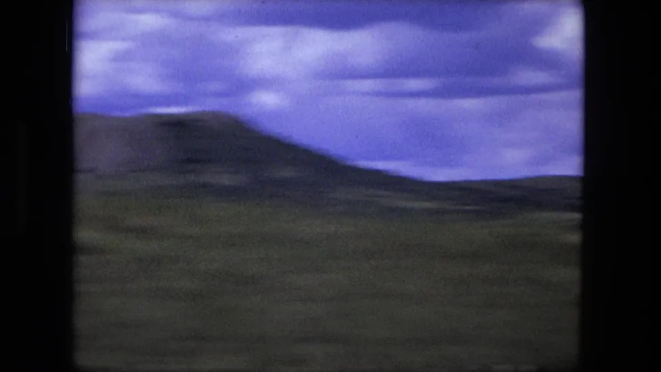 1976 8mm travel frame showing open scrubland, distant low hills, and a cloudy sky with visible film borders and color shift.